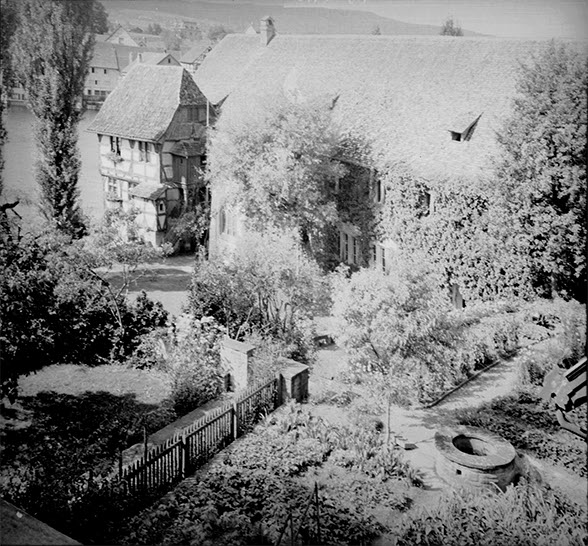 Blick von einem erhöhten Standort in einen Garten und auf Gebäude eines Klosters. Auf den Dächern liegen Biberschwanzziegel, die Mauern sind mit Efeu überzogen. Aufnahme in Schwarz/Weiss.